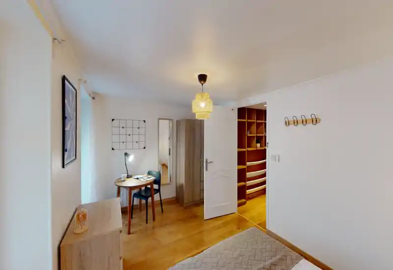 Small cozy bedroom with warm wooden flooring, a partially visible double bed, a compact desk and chair by the window, wardrobe and soft woven lighting—bright, well-styled and suitable for single or couple stays.