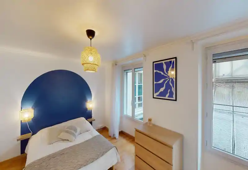 Bright, styled bedroom with a comfortable double bed, warm woven pendant and wall lamps, wooden floor and a blue painted headboard accent; two windows provide natural light and a small dresser offers storage.
