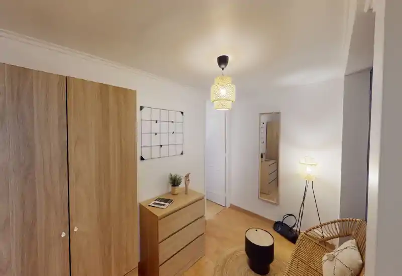 Cozy, well-styled small bedroom corner with natural-wood wardrobe, matching chest of drawers and wicker seating; warm lamps create a welcoming atmosphere.