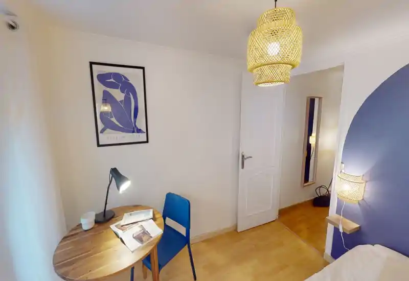 Compact bedroom corner with a small wooden table, reading lamp and a single bed visible at the frame's right edge; calm blue and white decor with warm woven lighting.