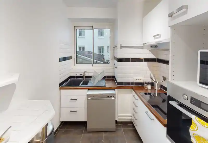Compact modern kitchen with white cabinets, wooden countertops, subway-tile backsplash and a window providing natural light. Includes sink, stovetop and dishwasher — efficient layout for a small apartment.