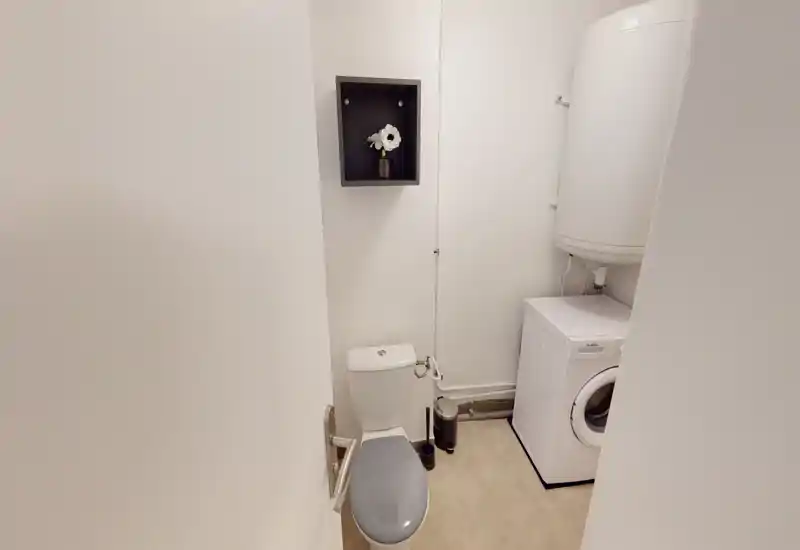 Compact bathroom with a toilet, a washing machine and a water heater; simple, clean white walls and good lighting.