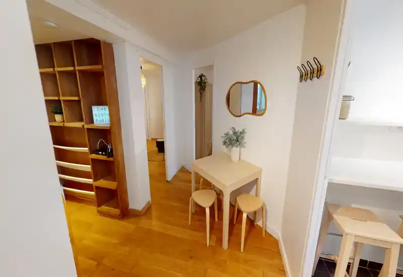 Compact entryway with wooden flooring, a small table and stools, wall hooks and shelving — bright and neatly styled for a welcoming arrival.