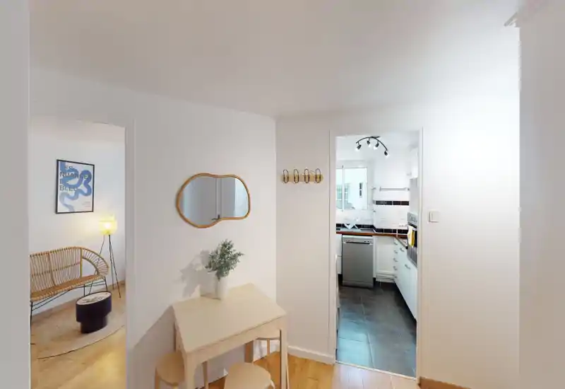 Bright, minimal entrance area with a small console table, mirror and coat hooks; view leads into a compact kitchen and adjoining living area.
