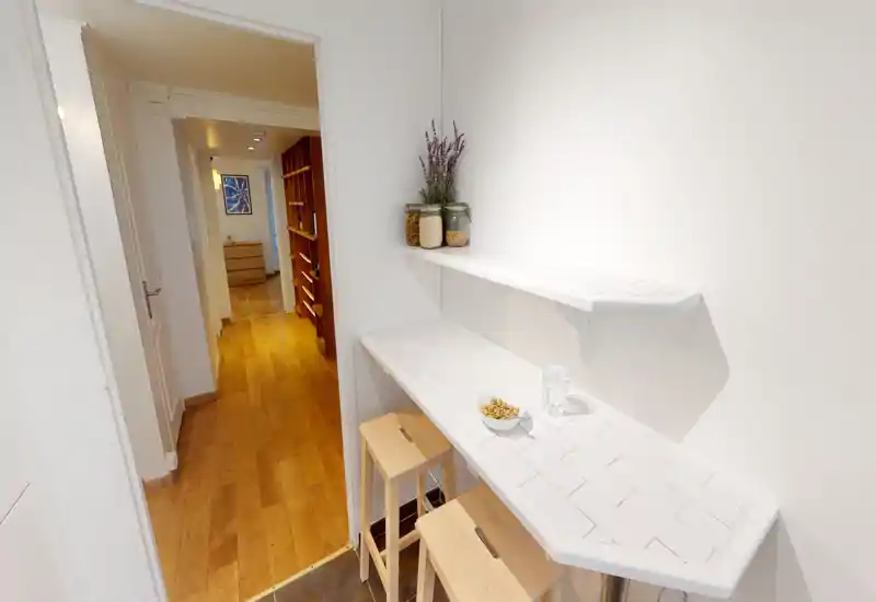 Compact kitchenette with a white tiled breakfast bar, two wooden stools, decorative jars and a bright corridor visible beyond — ideal for a studio or small apartment.
