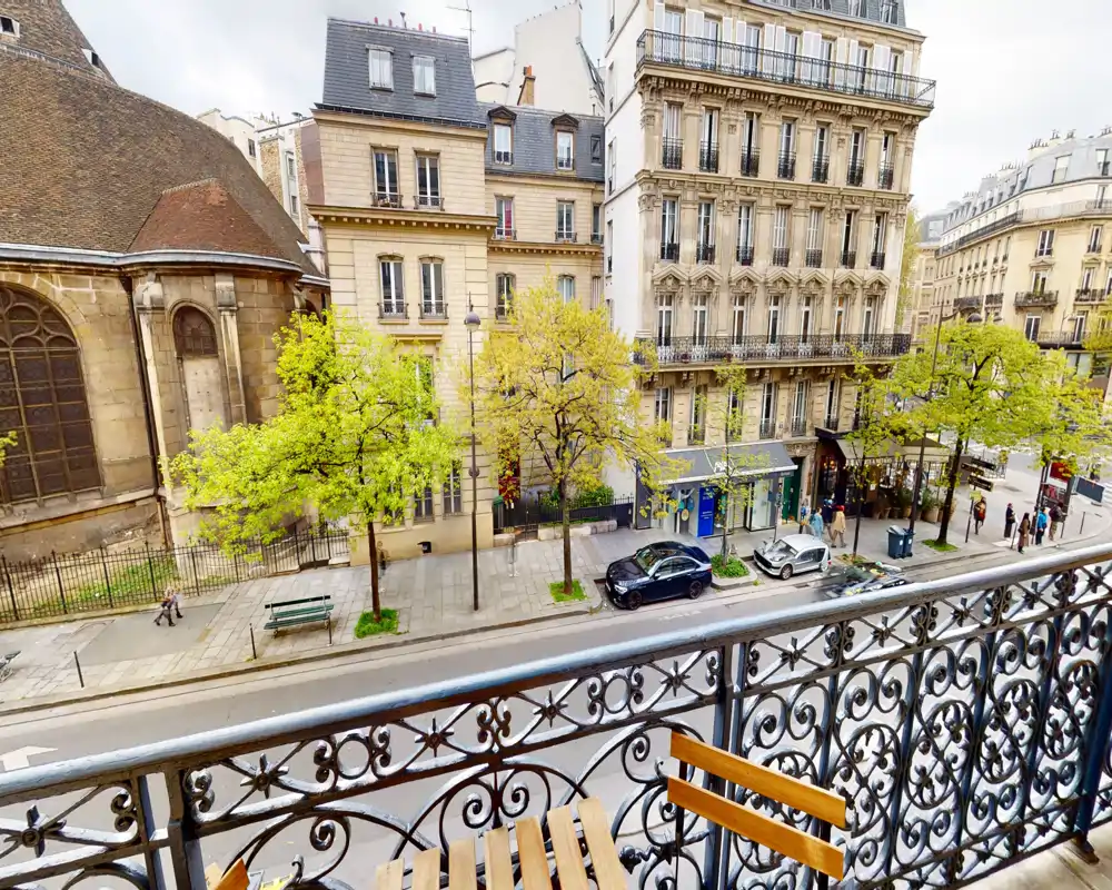 Sunny balcony view overlooking a Parisian street with classic Haussmann-style buildings, decorative iron railing and a small wooden bistro table.