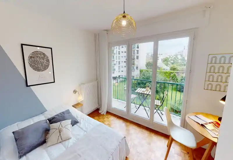 Bright, stylish bedroom with a comfortable double bed, decorative pillows and large glass doors opening to a small balcony with outdoor seating and garden views.
