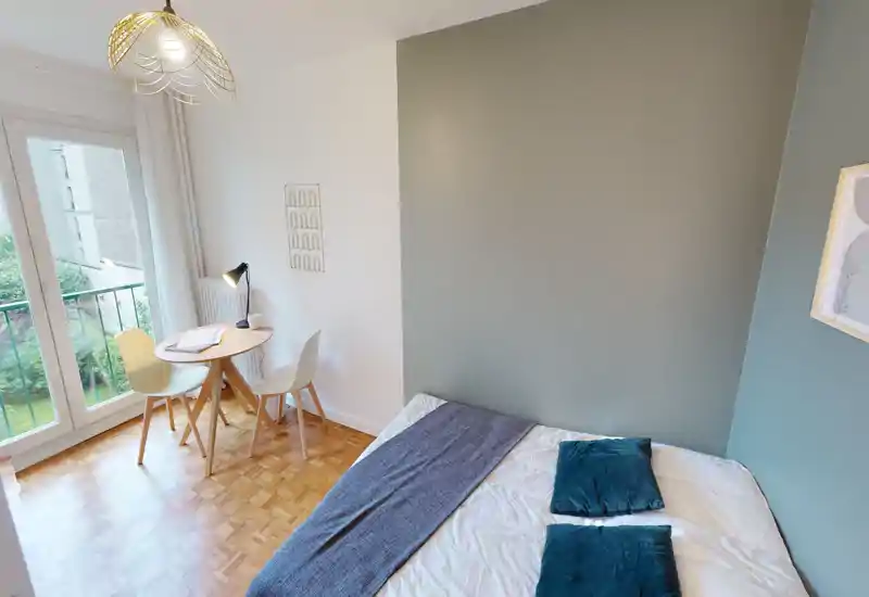 Bright, simply styled bedroom with a double bed against a muted accent wall, a small round table and chairs by the balcony door letting in natural light.
