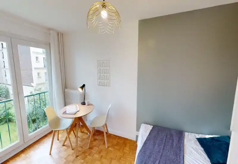 Compact, bright bedroom with a single bed, soft textiles and a small dining/work table next to a large window offering garden views.