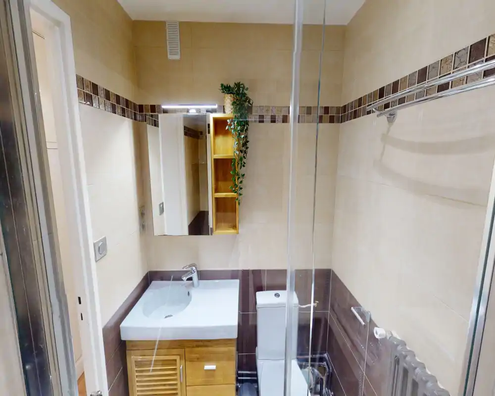 Modern compact bathroom with a vanity sink, mirror cabinet and toilet in warm beige and brown tiles; glass shower partition visible on the right.