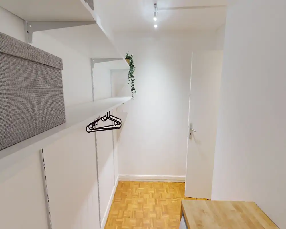 Small fitted storage/entry space with wall-mounted shelves, hangers and parquet flooring. Bright white walls and compact layout ideal for coats or a wardrobe niche.