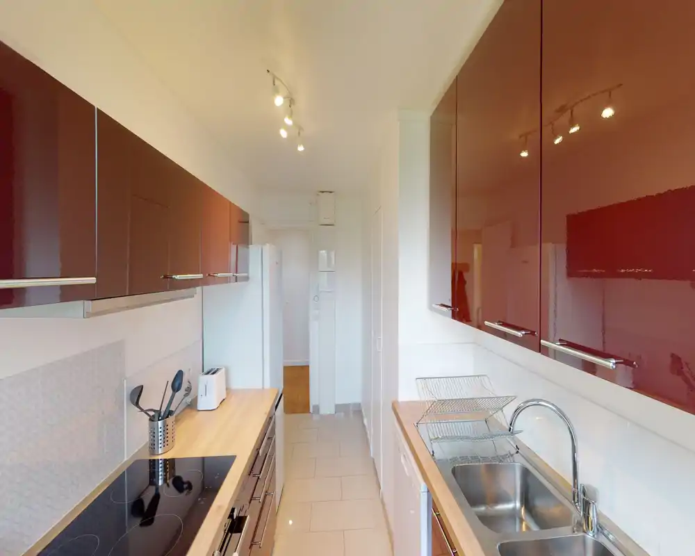 Modern galley kitchen with glossy burgundy cabinets, wood countertops, electric cooktop and stainless sink; bright, well-organized and suitable for meal preparation.