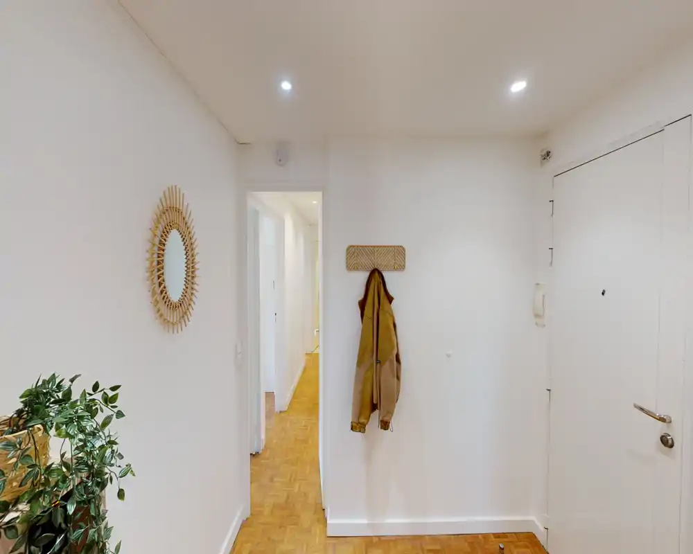 Bright, minimal entry hall with a coat hook, decorative mirror and parquet flooring — a welcoming entrance area leading to the interior corridor.