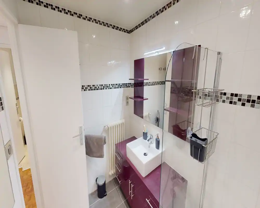Bright, modern bathroom with white tiled walls, a purple vanity and sink, mirror with integrated light, glass shower partition and chrome accessories.