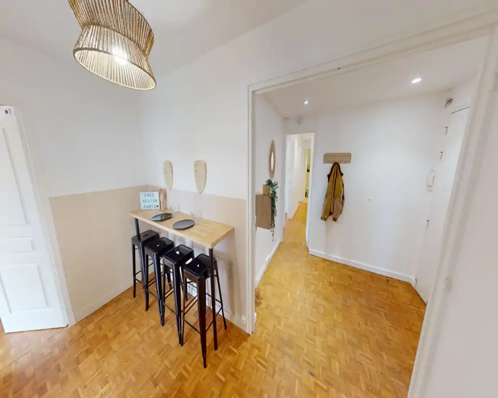 Compact dining area with a wooden high table and four black bar stools under a woven pendant lamp. Neutral two-tone walls and parquet flooring create a warm, minimalist atmosphere ideal for casual meals.