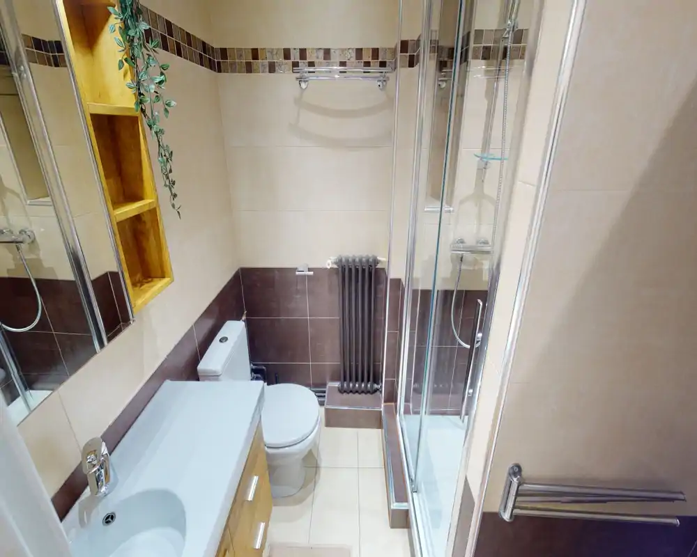 Compact, well-lit bathroom with a sink and vanity on the left, a toilet at center-left and a glass-enclosed shower on the right. Neutral beige and brown tiles and a wall-mounted radiator create a clean, modern look.