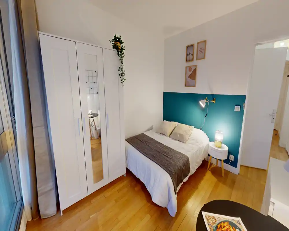 Chambre lumineuse et bien mise en scène avec un lit double contre un mur d’accent bleu sarcelle, table de chevet avec lampe et placard blanc. La lumière naturelle et la décoration donnent une ambiance chaleureuse.