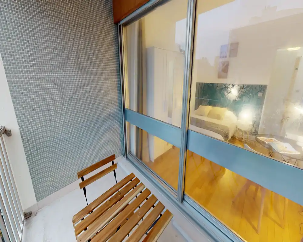 Petit balcon avec chaises pliantes en bois et mur carrelé, donnant sur une chambre à travers de grandes baies vitrées — idéal pour un café tranquille le matin.
