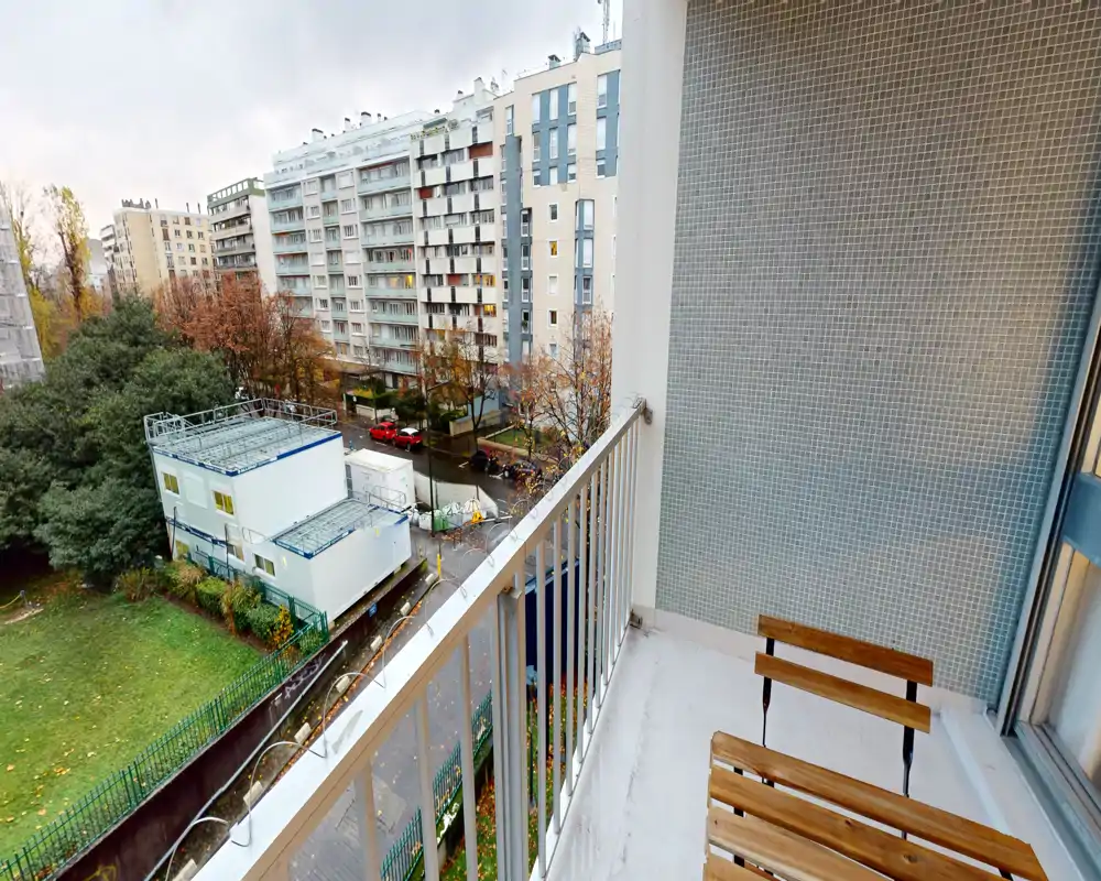 Balcon ensoleillé avec une chaise en bois simple, garde-corps métallique et vue sur la ville — idéal pour un café matinal face aux immeubles et au petit parc.