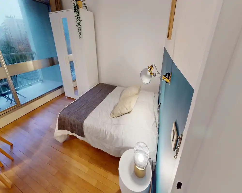 Chambre lumineuse et bien décorée avec lit double, lampe de chevet et armoire ; grande fenêtre et sol en bois pour une ambiance chaleureuse et aérée.