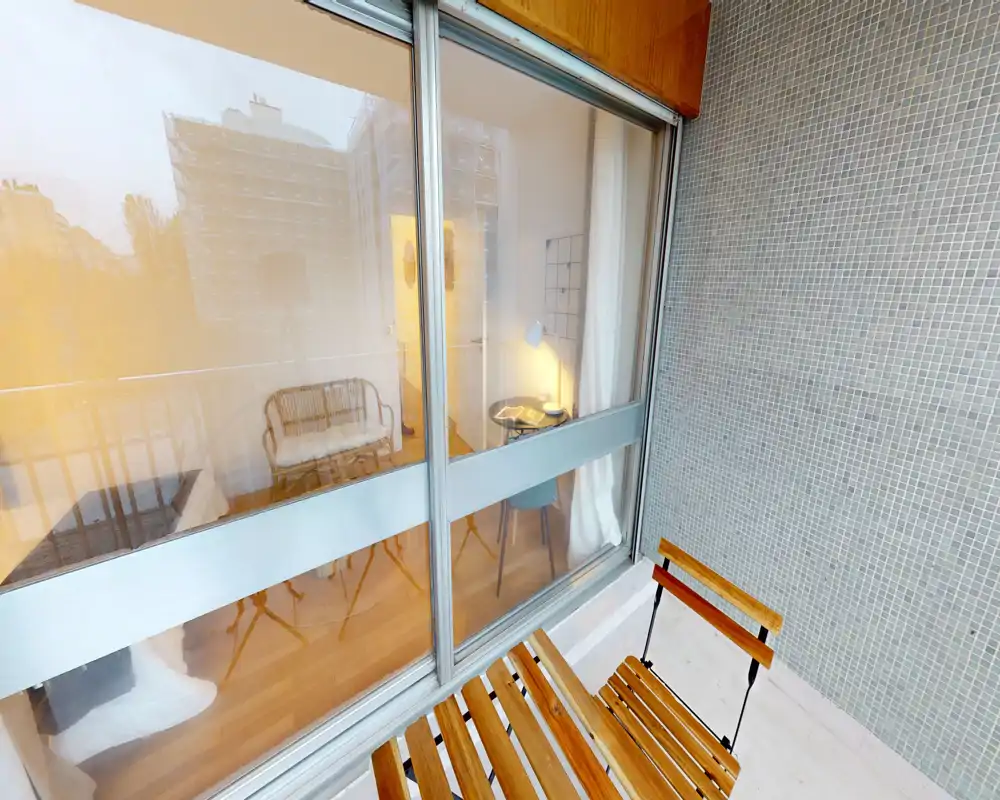Petit balcon avec chaises en bois à lattes et vue sur la pièce adjacente à travers de grandes baies vitrées coulissantes ; éclairage chaud agréable et mur carrelé.