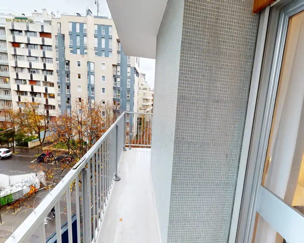 Balcon étroit avec garde-corps en métal donnant sur une rue résidentielle et des immeubles voisins ; idéal pour de petites chaises ou des plantes en pot.
