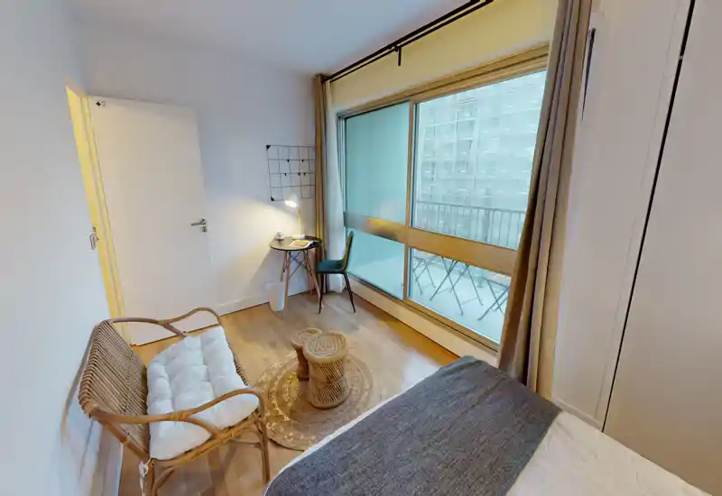 Bright, modern bedroom with a comfortable bed, wicker seating and a work nook by the large window that opens to a small balcony.