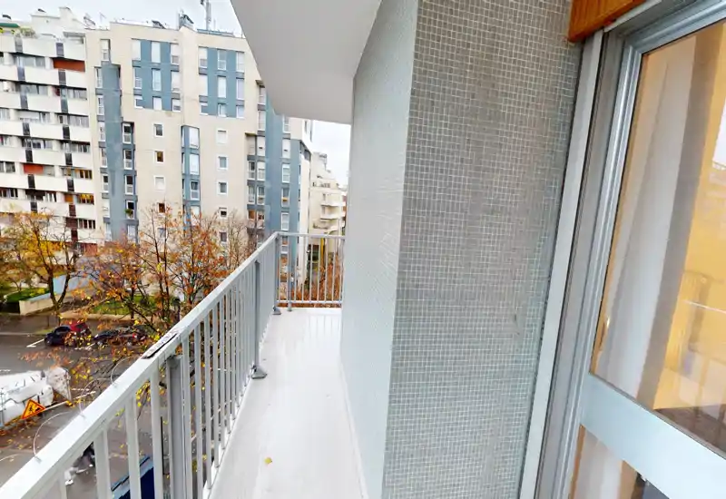 Sunny narrow balcony with metal railing, tiled wall and sliding glass door opening to the living area; overlooks residential buildings and street below.