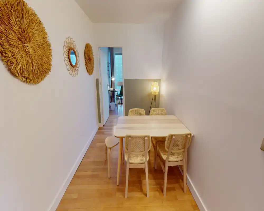Coin repas lumineux et compact avec table en bois et quatre chaises à dossier tressé, lampe d'ambiance chaude et décorations murales apportant une atmosphère cosy.