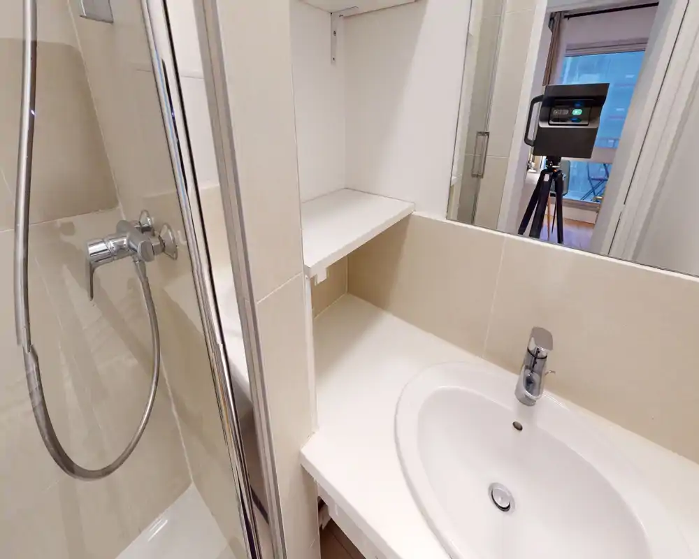 Salle de bains compacte et moderne avec lavabo blanc, robinet chromé et douche visible à gauche ; carrelage clair et miroir mural donnent un style propre et contemporain.