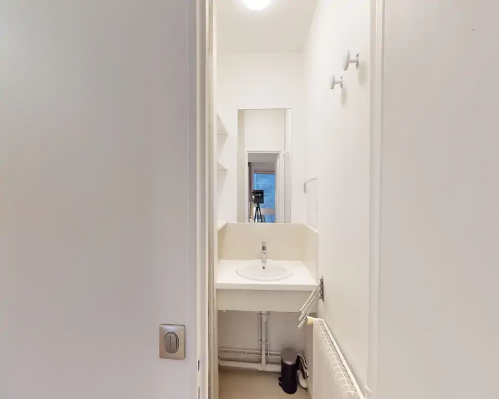 Petite salle de bain lumineuse avec un lavabo rond, robinet monocommande, miroir et tuyauterie apparente sous le plan; murs blancs pour une esthétique minimaliste.