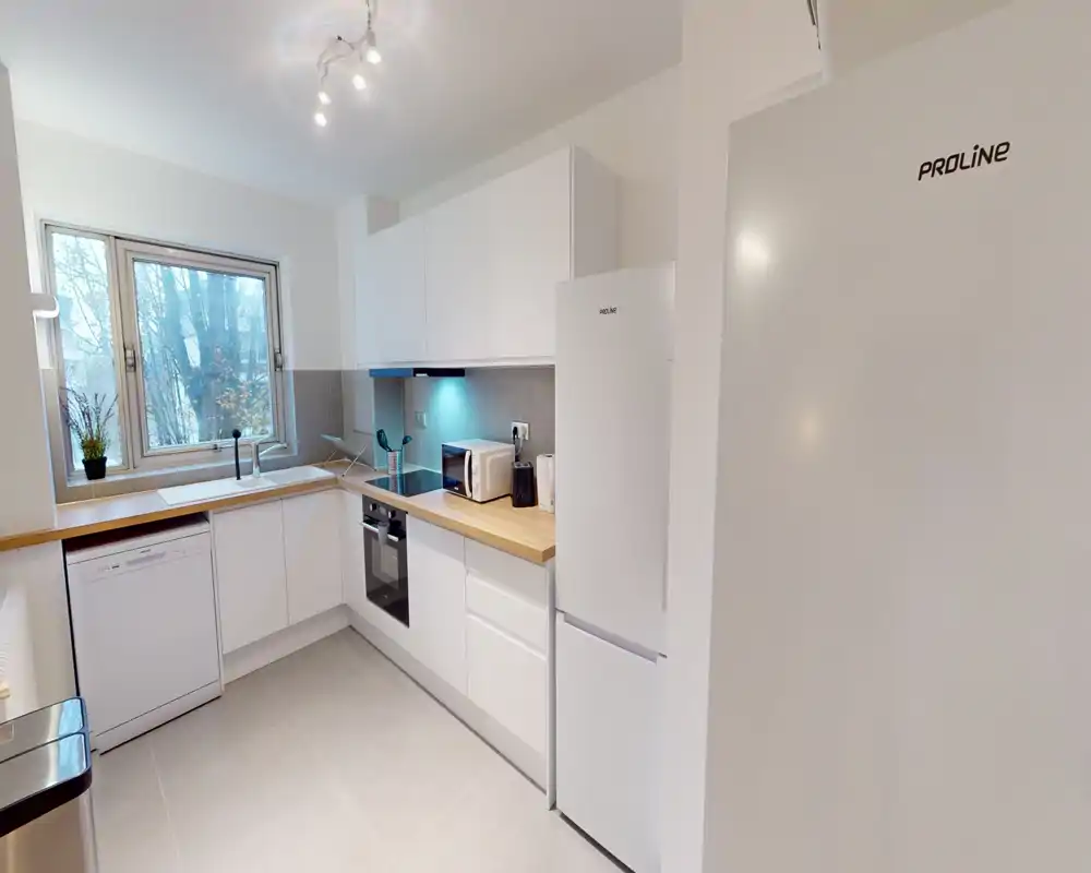 Cuisine moderne équipée avec placards blancs, plan de travail en bois, four encastré, réfrigérateur et lave-vaisselle; grande fenêtre apportant une belle lumière naturelle, espace propre et fonctionnel.