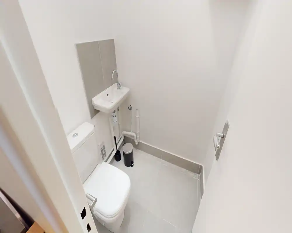 Salle de bains compacte et bien éclairée avec WC, petit lavabo mural et plomberie apparente, sol en carreaux gris clair.