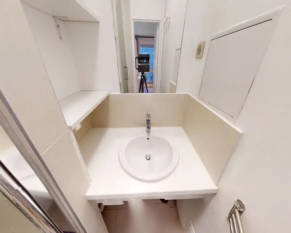 Petit coin lavabo de salle de bain avec vasque ronde encastrée, mitigeur monocommande, étagère intégrée à gauche et miroir reflétant l'entrée. Surfaces blanches et carrelage neutre pour un espace de toilette simple et fonctionnel.