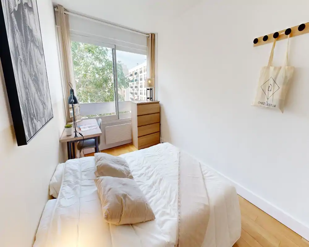 Chambre lumineuse et minimaliste avec lit double, oreillers douillets et sol en bois ; grande fenêtre apportant une belle lumière naturelle et un petit bureau près de la fenêtre.