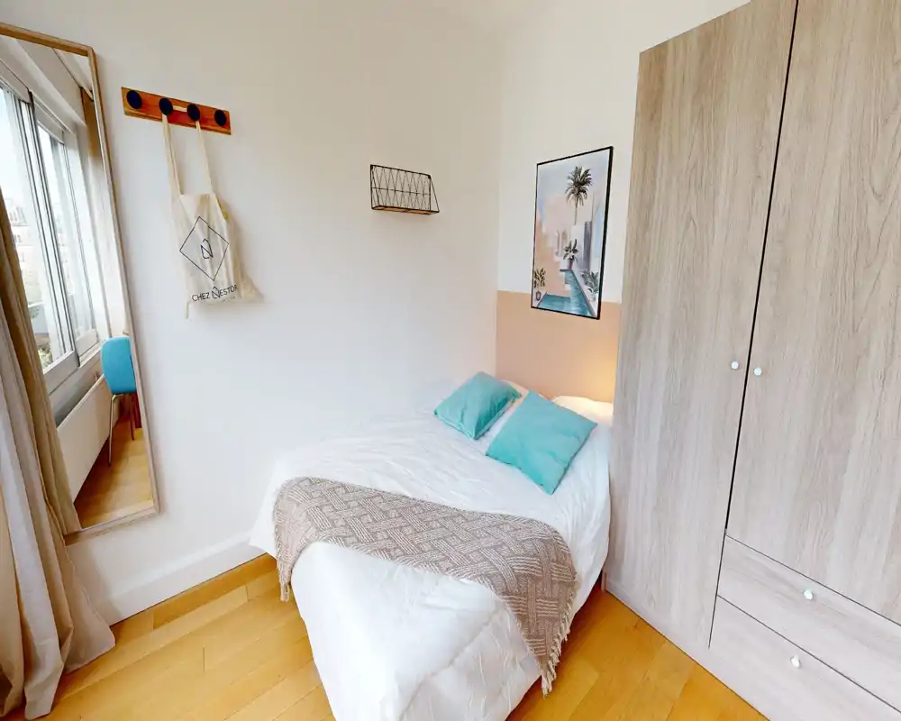Chambre lumineuse et bien mise en scène avec un lit double en linge blanc, deux coussins turquoise et un plaid texturé; grande armoire et fenêtre avec miroir complètent l'espace.