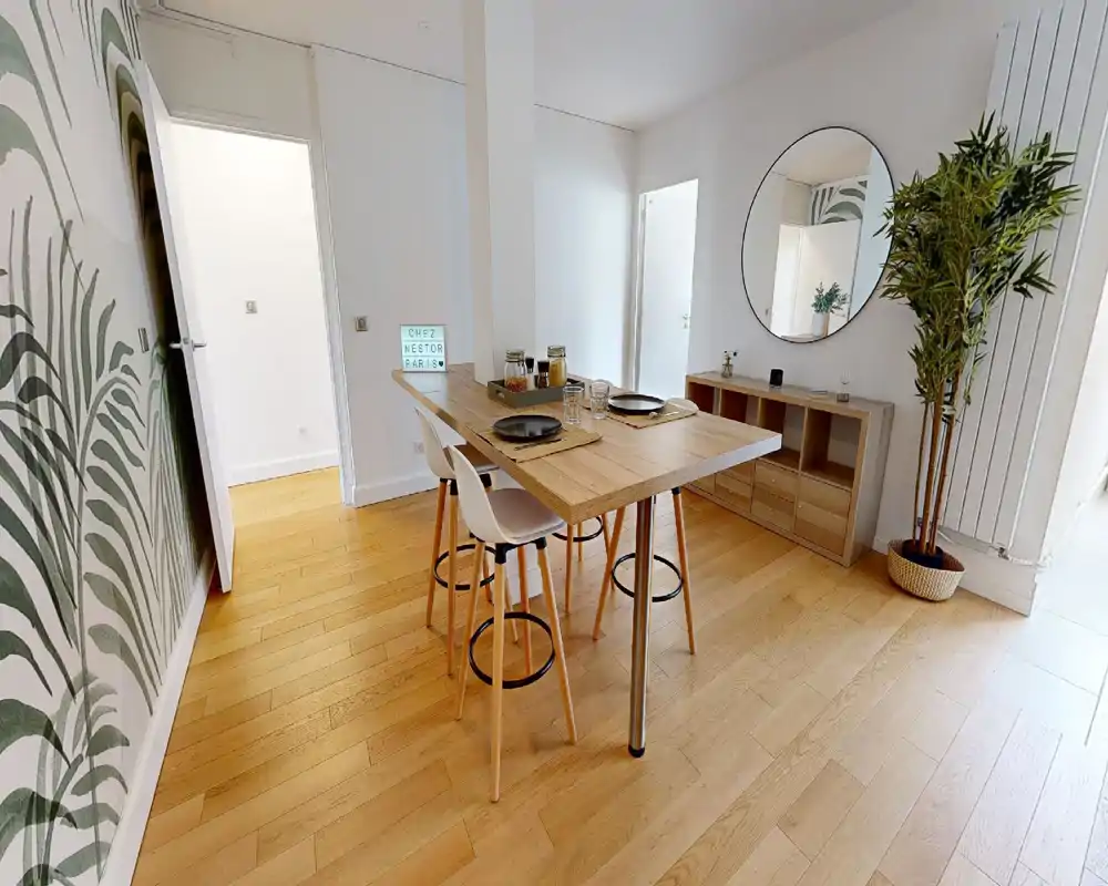 Coin repas moderne et lumineux avec une grande table haute en bois pour deux, des tabourets et des plantes décoratives. Sol en bois chaleureux, grand miroir rond et décoration contemporaine épurée.