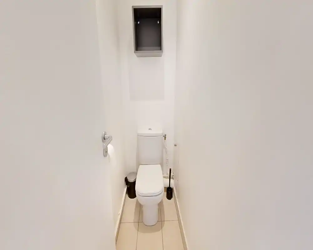 WC compact et propre avec murs blancs, sol carrelé, toilette, brosse et petite poubelle ; cabinet lumineux et sobre.