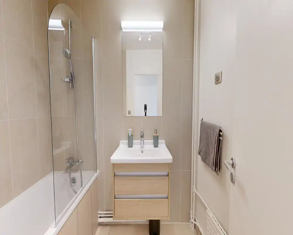 Salle de bains moderne avec baignoire et pare-douche en verre, meuble-lavabo mural, miroir et porte-serviettes ; carreaux neutres et éclairage lumineux et uniforme.