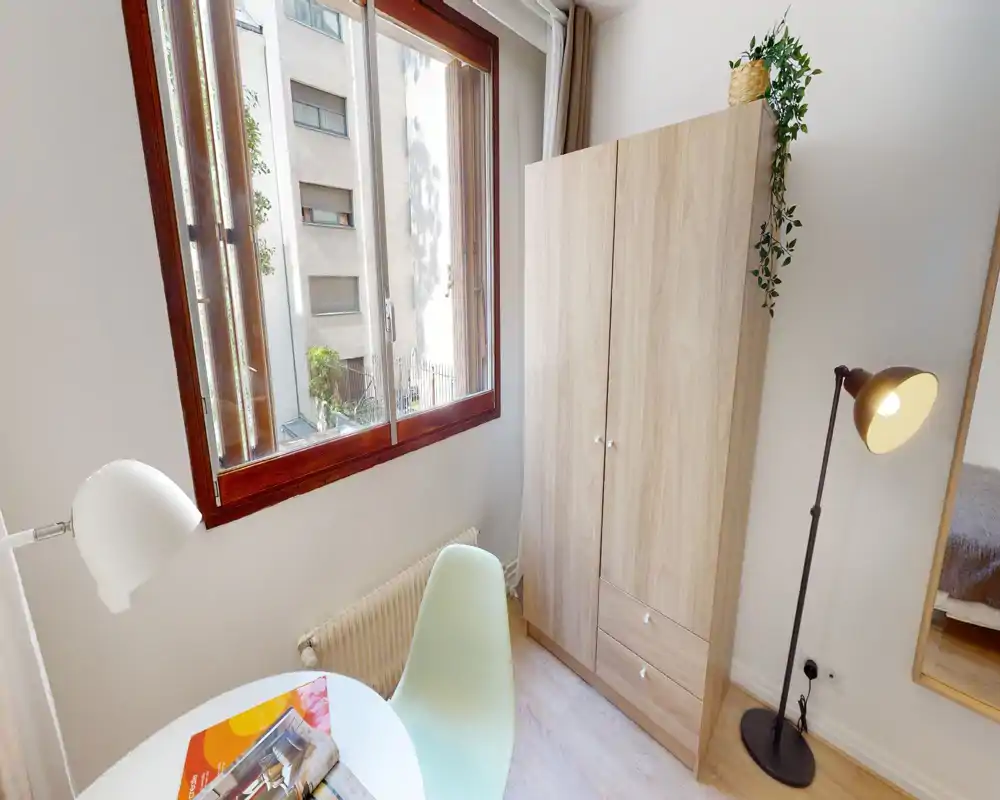 Petit coin de chambre lumineux avec une armoire, une lampe sur pied et une fenêtre laissant entrer beaucoup de lumière naturelle ; une partie du lit est visible sur le bord droit. Mobilier moderne et propre, atmosphère chaleureuse grâce à la lampe et à la plante.