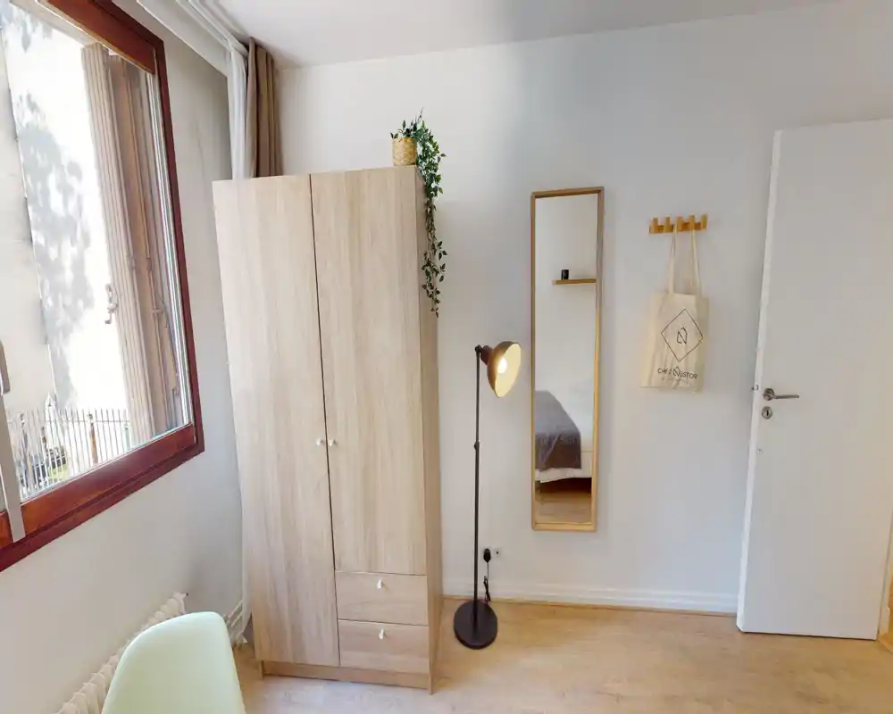 Coin de la chambre compact et bien éclairé avec une armoire en bois, une lampe sur pied et une glace pleine longueur reflétant une partie du lit. Décor neutre et lumière naturelle d'une grande fenêtre pour une ambiance calme et accueillante.