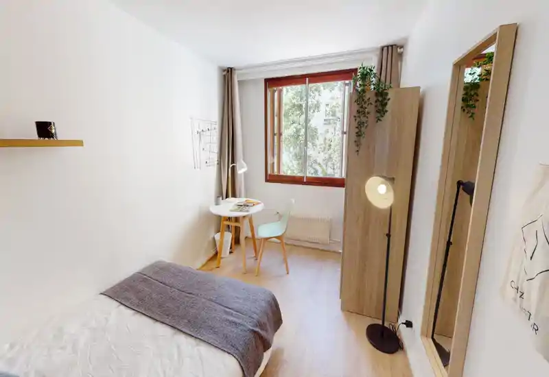 Chambre lumineuse et compacte avec un lit simple, une petite table ronde près de la fenêtre, une armoire, une lampe sur pied et un miroir en pied — parfaite pour étudiant ou locataire seul.