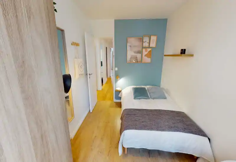 Chambre moderne et lumineuse avec un lit double contre un mur bleu pastel, cadres décoratifs et une petite étagère flottante. Parquet et textiles chaleureux donnent une ambiance confortable, idéale pour la location.