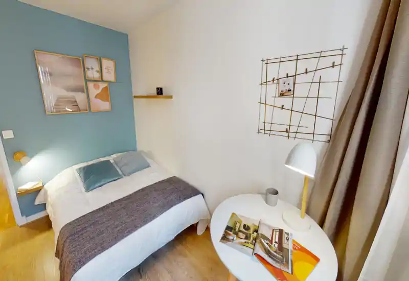 Chambre lumineuse et bien décorée avec un lit double contre un mur bleu, tableaux décoratifs, lampe de chevet et petite table ronde — chaleureuse et moderne.