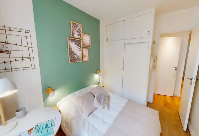 Chambre chaleureuse et lumineuse avec lit double en linge neutre, coussins décoratifs et tableaux sur un mur vert; placard intégré et sol en bois chaleureux.