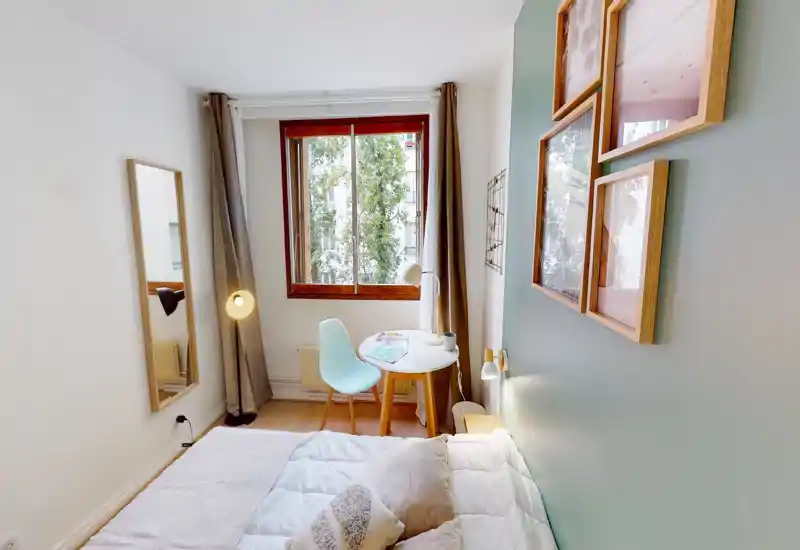 Chambre lumineuse et chaleureuse avec lit bien fait au premier plan, petit bureau rond près d'une grande fenêtre et cadres décoratifs sur un mur vert pâle.