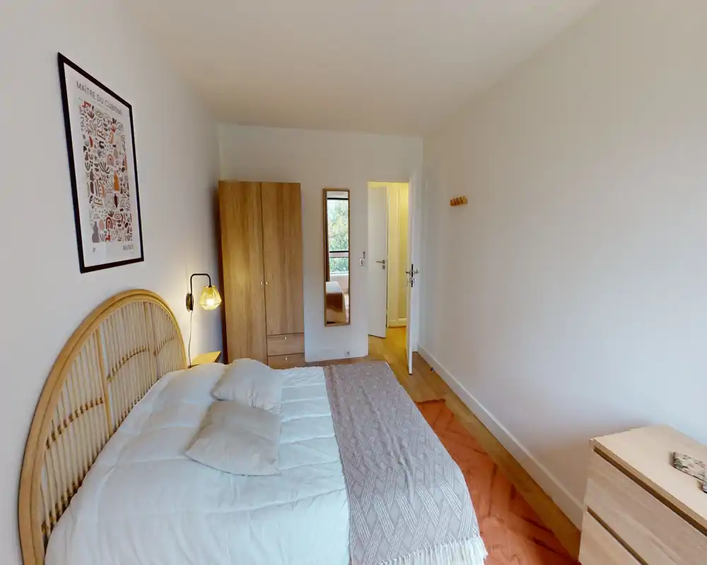 Chambre lumineuse et soignée avec tête de lit en rotin, lit double en literie blanche, lampe de chevet et armoire; tons neutres et lumière naturelle pour une atmosphère chaleureuse.