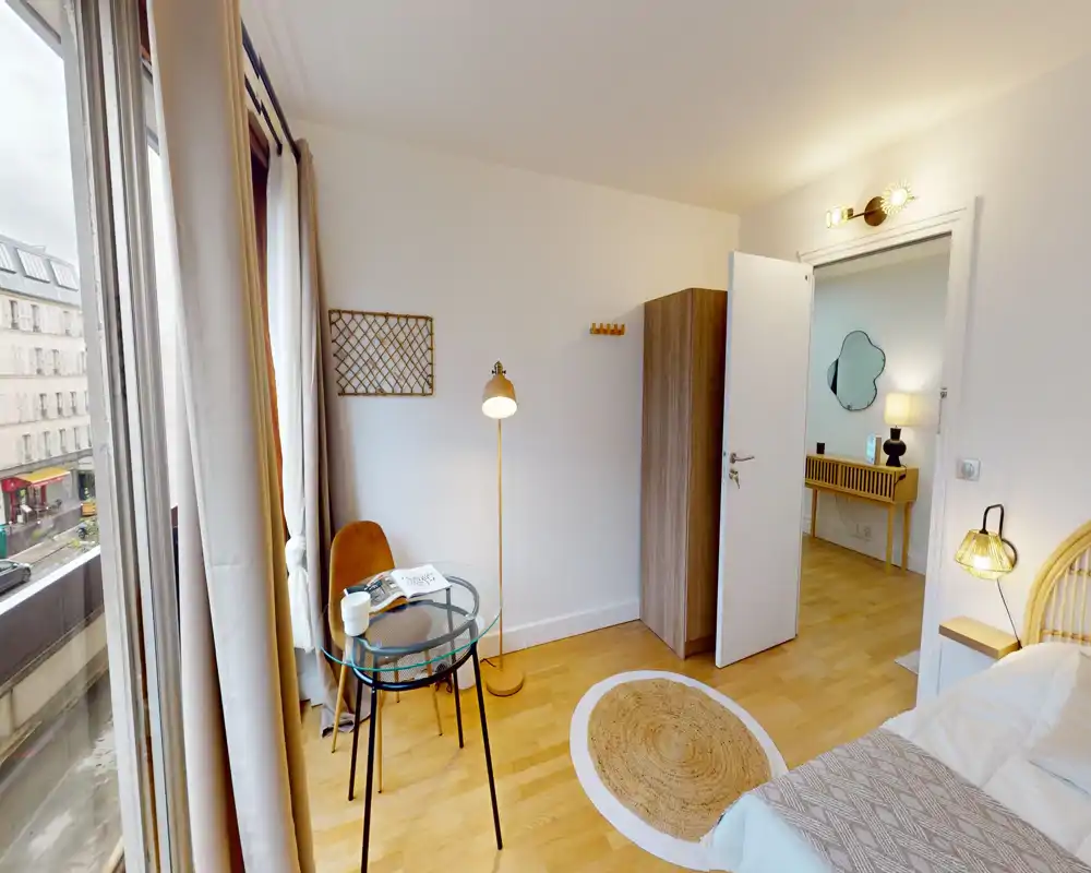 Bright, cozy bedroom corner with a partial view of the bed, a round woven rug, a small glass table with chair and a tall window letting in abundant natural light.