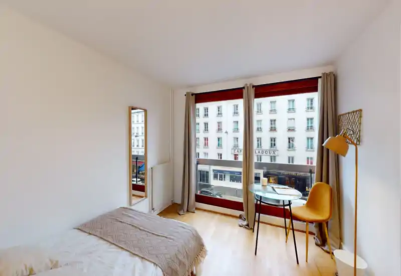 Bright, modern bedroom with a comfortable bed, large street-facing window and a cosy reading nook with a small round table and chair.