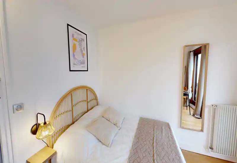 Bright, cozy bedroom with a rattan headboard, bedside lamp and neatly made double bed. The room features warm wooden flooring and a full-length mirror reflecting an adjacent area.